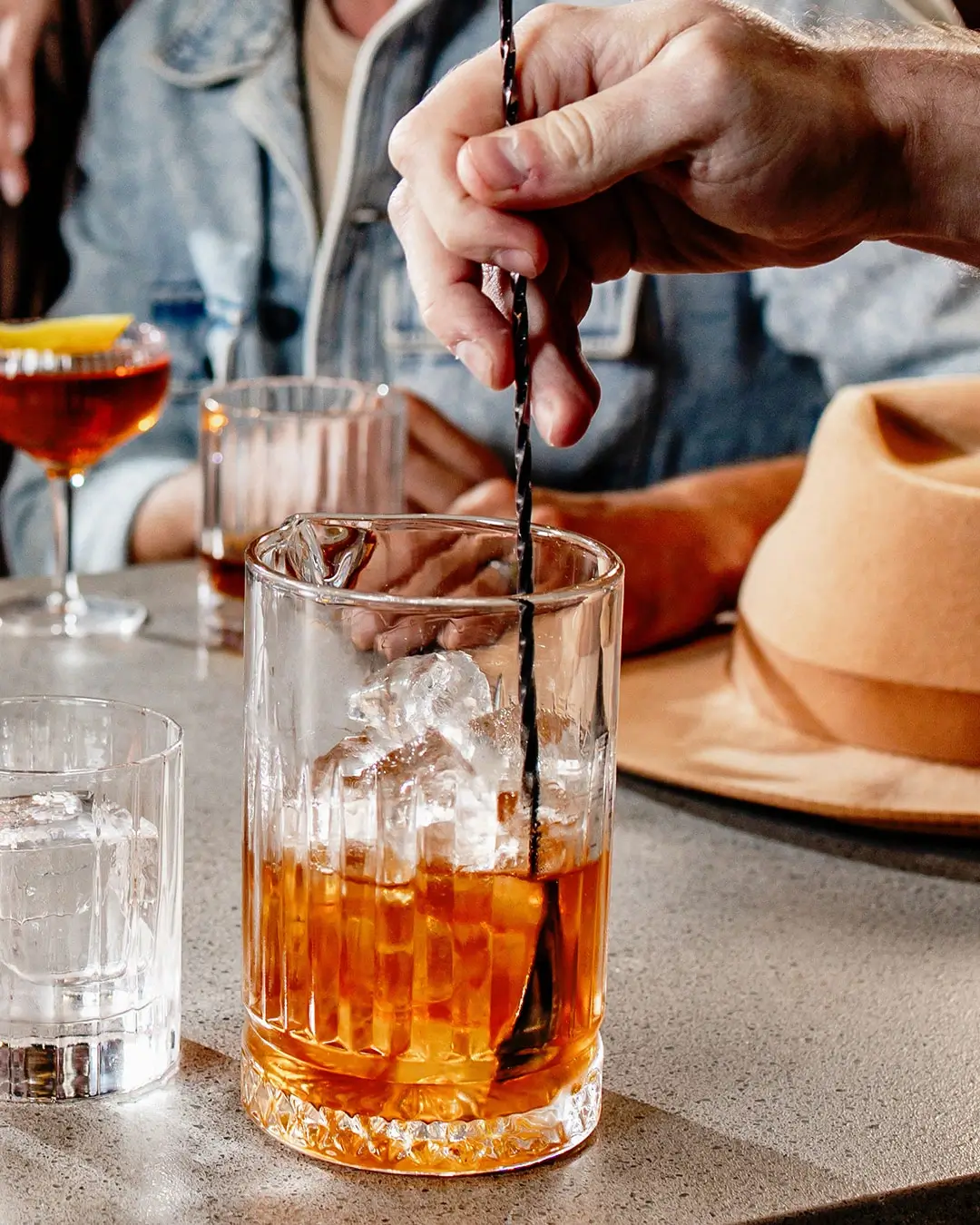 Hand stirring whiskey cocktail with ice and bar spoon in rocks glass at bar counter