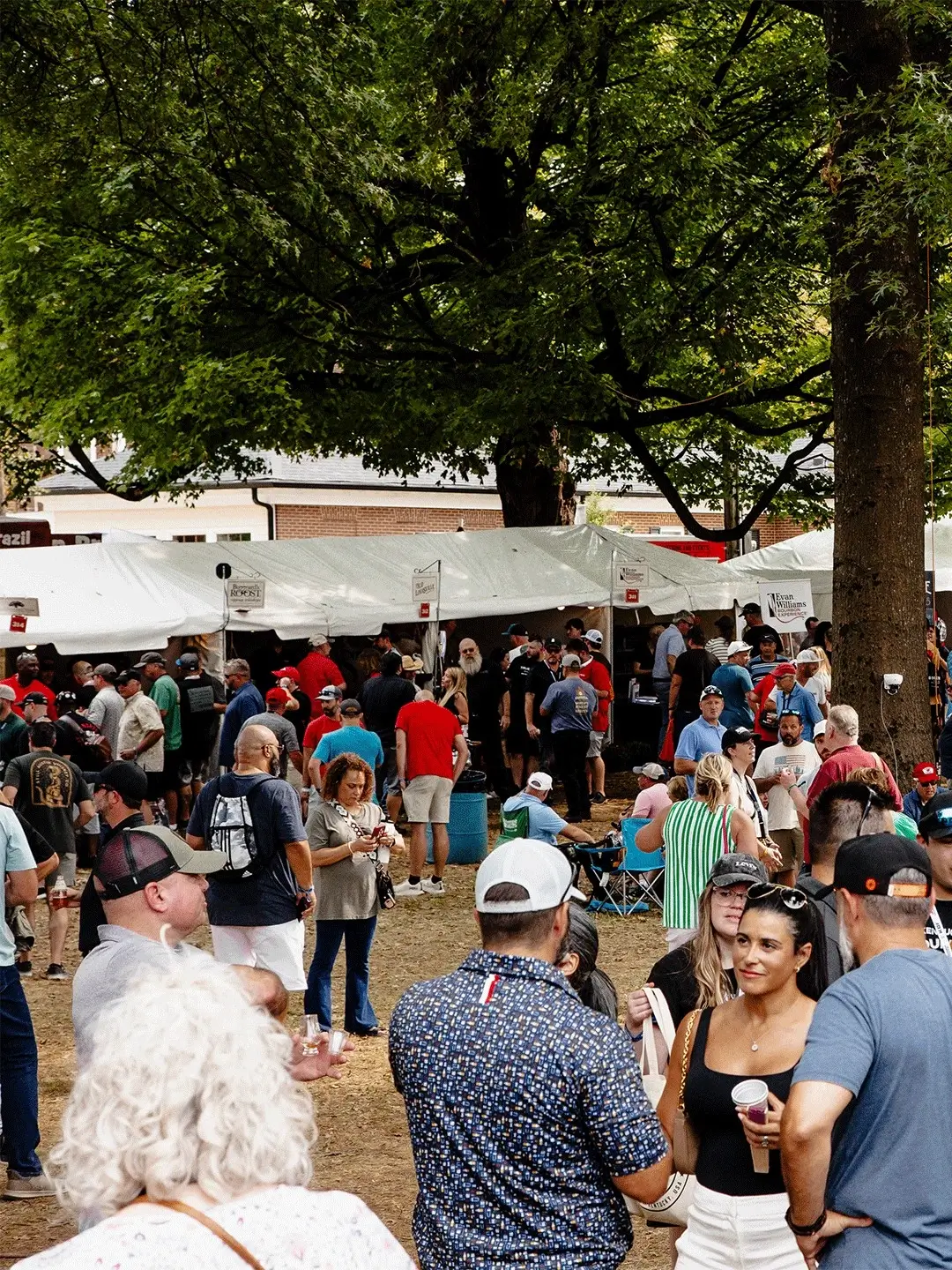 Crowds of people mingling at an outdoor bourbon festival with white tents and vendor booths beneath large trees