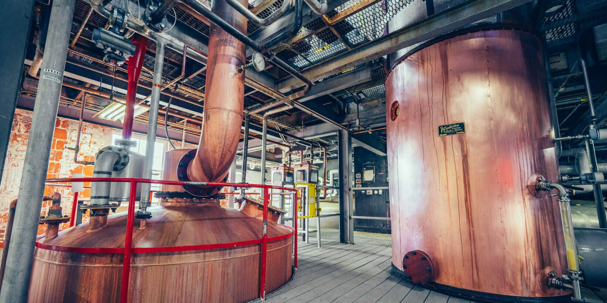 Large copper stills and fermentation tanks with red railings inside an industrial distillery or brewery facility