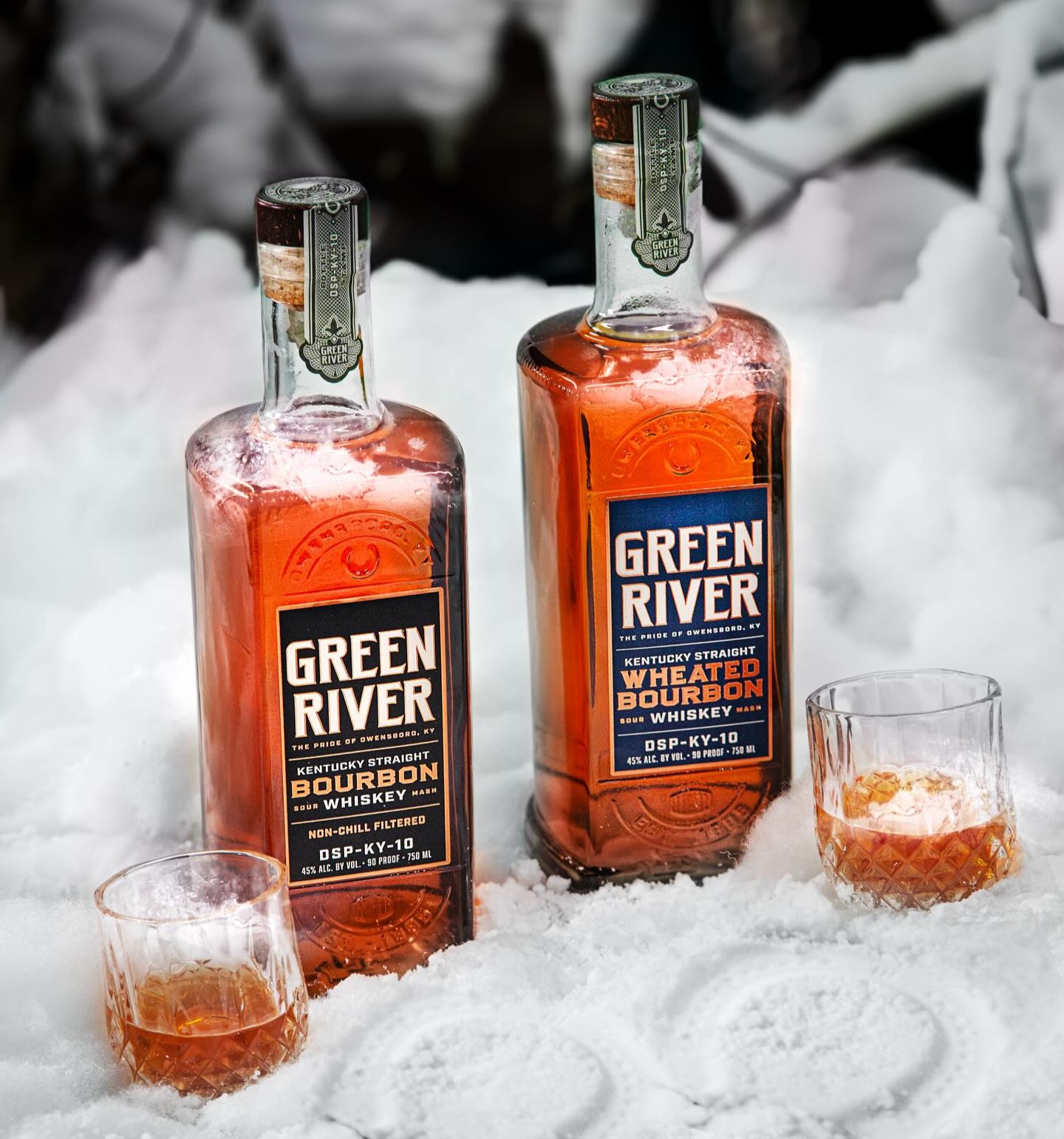 Two Green River bourbon whiskey bottles in snow with two glasses of whiskey on ice